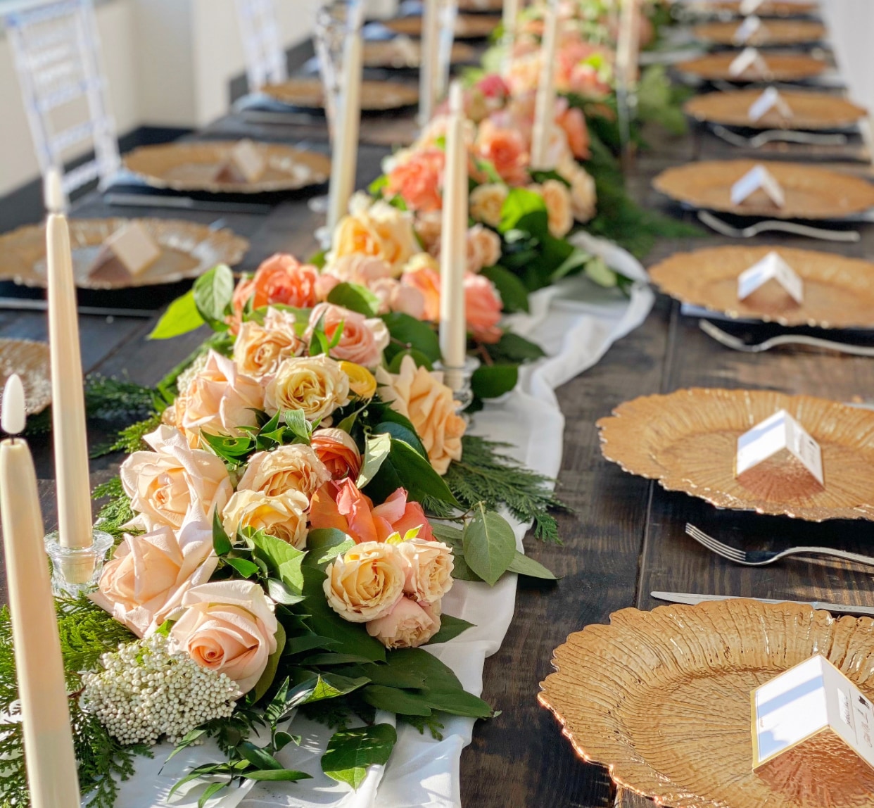 a long head table with a table runner made of fresh flowers in creme, orange and coral color tones, with flowers like roses, ranunculus and greeneries. the table is also accented with crystal candlesticks with led taper candles.